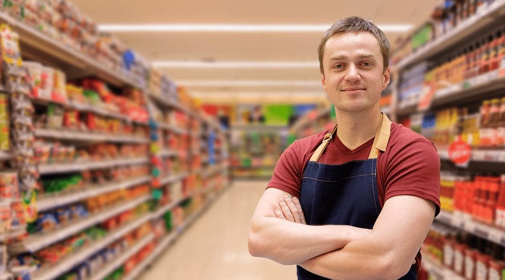 Smiling store employee with arms crossed, promoting products in a supermarket aisle. Product Promoter App.