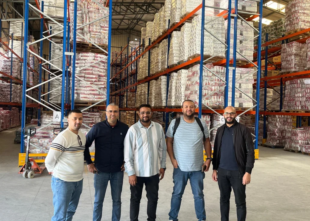 Five men stand in a large warehouse filled with stacked pallets of products.  Industries.