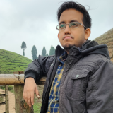 Headshot of a young man in glasses and a dark jacket, leaning on a wooden fence with a tea plantation in the background.