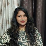 Headshot of a woman with long dark hair, wearing a beige and brown patterned top, against a curtain backdrop.